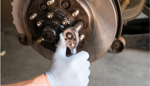 Close-up of a mechanic's gloved hand using a ratchet wrench to work on a car's brake rotor and hub assembly. The brake components are clearly visible, with the mechanic tightening or loosening a bolt on the vehicle in a garage setting.