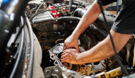 A mechanic working under the hood of a car inside a garage. The mechanic is focused on inspecting or repairing the engine, surrounded by various automotive tools and equipment in the background.